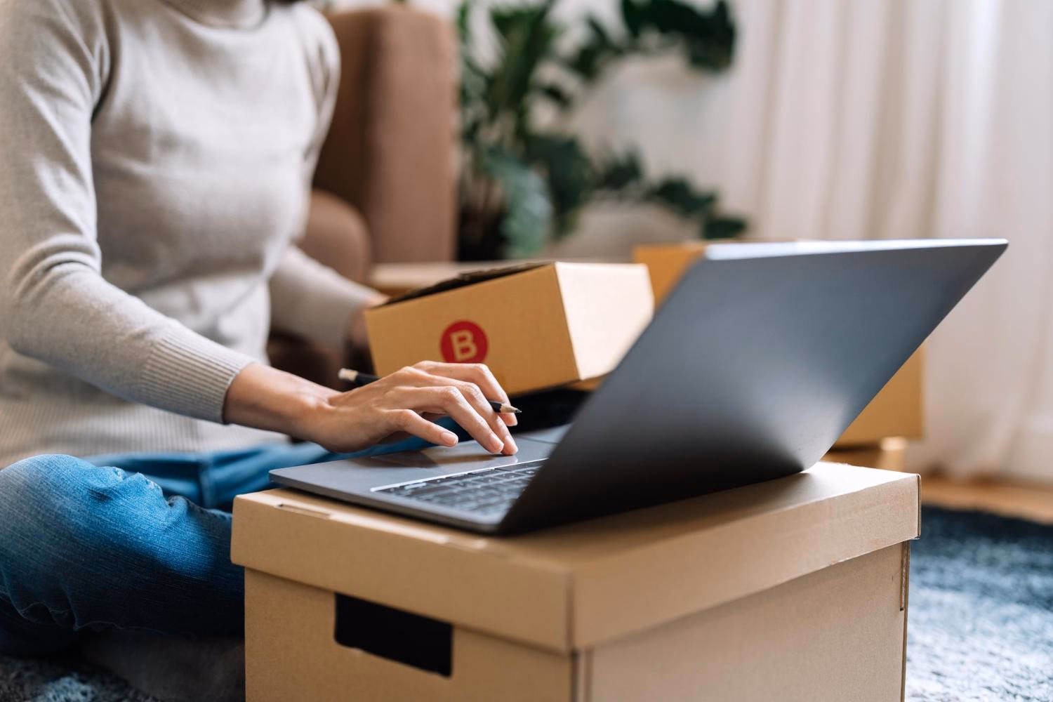 Kobieta siedząca na podłodze z laptopem na kartonie i paczkami kurierskimi obok, prowadzi biznes online w domu