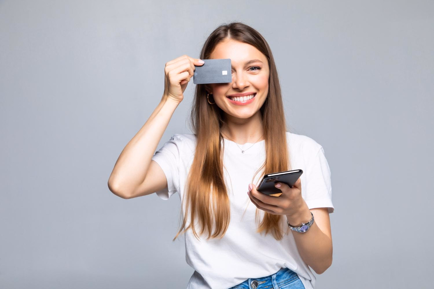 Uśmiechnięta młoda kobieta w białej koszulce trzyma kartę kredytową przy oku i smartfon w dłoni