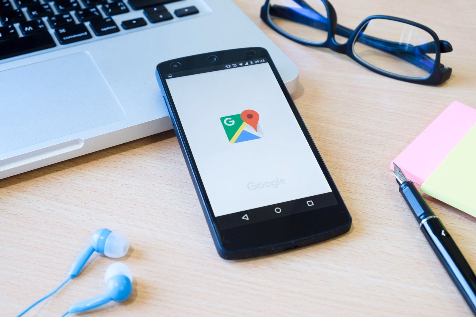 Smartfon z uruchomioną aplikacją Google Maps leżący na biurku obok laptopa, słuchawek i okularów